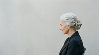 Senior woman with gray hair in updo hairstyle, wearing black blazer, smiling in profile against neutral background. Suitable for business, corporate, and lifestyle editorial use.