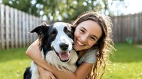 Young woman with curly brown hair embracing border collie dog outdoors in backyard garden, both smiling at camera showing happiness and companionship on sunny day.