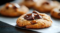 Fresh baked chocolate chip cookie with melted dark chocolate chunks on textured surface, selective focus on gooey chocolate pieces, macro food photography.