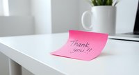 Pink sticky note with thank you message on white desk in modern office workspace with plant and laptop for gratitude and appreciation concepts.