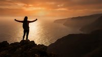 Silhouette of person with raised arms celebrating success on mountain cliff overlooking ocean at golden sunset. Freedom, achievement, motivation concept.