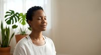 Peaceful African American woman with closed eyes practicing mindfulness meditation at home with green plants creating serene wellness atmosphere.