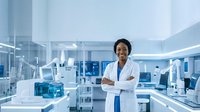 African American female scientist in white lab coat standing confidently with crossed arms in modern research laboratory with advanced equipment and blue lighting.