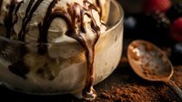 Vanilla ice cream topped with chocolate sauce drizzle, cocoa powder, and wooden spoon on dark surface for dessert and food styling.