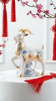 Porcelain goat figurine with gold accents and blue floral patterns, surrounded by red tassels and pink flowering branches on white background. Lunar new year decoration.
