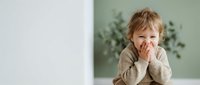 Adorable toddler girl with blonde hair covering face with hands in playful peek-a-boo gesture, sitting indoors against modern minimalist background.