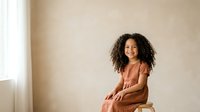 Young woman with curly dark hair and medium skin tone wearing rust-colored dress, sitting on wooden stool against neutral beige wall with natural light from window, suitable for lifestyle and portrait projects.