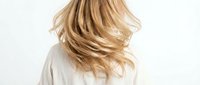 Young woman with flowing blonde hair tossing her head, wearing a white shirt, captured from behind on a light background.