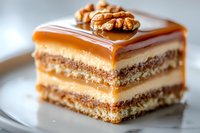 Layered caramel cake slice with walnut topping on white plate, featuring alternating layers of moist sponge, cream filling and glossy caramel sauce garnish.