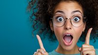 Young African American woman with curly hair and glasses showing surprised expression with open mouth and raised fingers against turquoise background.