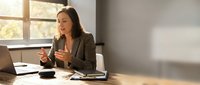 Professional businesswoman using smartphone at modern office desk with laptop and coffee cup during golden hour lighting for corporate communications.