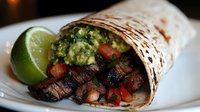 Grilled steak burrito with fresh guacamole, diced tomatoes and lime wedge on white plate. Mexican cuisine wrap filled with seasoned beef and vegetables.