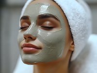 Young woman with clay facial mask and white towel on head enjoying spa treatment and skincare routine at wellness center, closeup portrait.