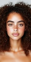 Close-up portrait of young mixed race woman with natural curly brown hair, freckles, and brown eyes looking directly at camera against light background.