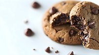 Freshly baked chocolate chip cookie broken in half showing soft gooey center with scattered chocolate chips on white surface, macro food photography.