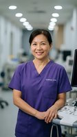 Mature Asian female healthcare professional wearing purple scrubs with clinical lead designation standing confidently in modern medical facility with equipment and overhead lighting, suitable for healthcare, medical services, and hospital staff recruitment materials.