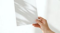 Close-up of a hand holding a blank white paper sheet against a white wall with natural shadow overlay, suitable for mockups and presentations.