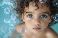 Close up portrait of young mixed race child with curly hair and bright blue eyes surrounded by soap bubbles against turquoise background, dreamy atmosphere.