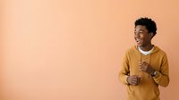 Young African American man wearing mustard yellow hoodie with white undershirt, smiling and gesturing with thumbs up against warm peach background, suitable for lifestyle, motivation, and positive messaging campaigns.