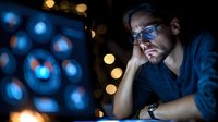 Focused man with glasses working late at computer screen with data visualization, illuminated by blue light in dark office environment.