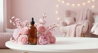 Essential oil bottle with pink roses and cherry blossoms on white table in cozy bedroom interior with fairy lights for aromatherapy and wellness.