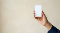 Hand holding blank white rectangular card against neutral beige background, ready for mockup designs, branding presentations, business cards, or promotional materials.