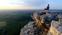 Young Asian woman in meditation pose sits on dramatic cliff edge overlooking vast green forest landscape during golden sunset hour, expressing serenity and mindfulness.