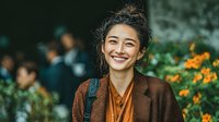 Young Asian woman with curly hair smiling joyfully in autumn setting with orange flowers, wearing brown coat and backpack for lifestyle and travel content.