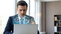 Professional businessman analyzing digital data interface with holographic technology display while working on laptop in modern office environment.