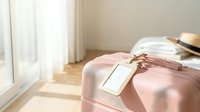 Pink suitcase with luggage tag ready for travel vacation trip in bright modern bedroom with natural sunlight streaming through window curtains.