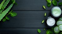 Fresh aloe vera leaves with mint and cucumber slices around cosmetic cream jars on dark wooden background for natural skincare and beauty treatments.