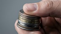 Close-up of a hand holding a stack of mixed currency coins, showing various denominations and metallic finishes against a neutral gray background. Use for finance, savings, investment, and wealth-related projects.