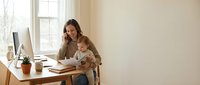 Young mother working from home office while holding baby, multitasking professional woman balancing career and childcare in modern workspace.