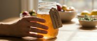 Hands holding glass jar of golden honey with organic label on wooden table with fresh fruits in warm kitchen setting for natural food concepts.