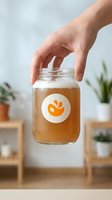 Hand reaching for glass jar with orange liquid and circular label in modern kitchen setting with plants and wooden shelves in soft natural lighting.
