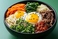 Korean bibimbap bowl with sunny side up eggs, minced beef, carrots, spinach, rice noodles, green onions and spicy sauce served on green background.