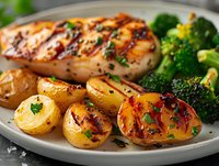 Grilled chicken breast with honey glaze served alongside roasted baby potatoes and steamed broccoli on gray plate, garnished with fresh herbs and black pepper.
