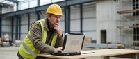 Senior construction worker in safety vest and hard hat using laptop computer at industrial warehouse site for project management and planning.