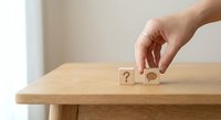 Hand placing wooden cube with question mark next to magnifying glass symbol on natural wood table representing problem solving and search concepts.