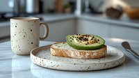 Fresh avocado toast on rustic sourdough bread served on speckled ceramic plate with matching coffee mug, healthy breakfast setting in modern kitchen.