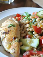 Grilled chicken breast with herb seasoning served alongside fresh chickpea salad with cucumber, tomato, and green onion on white plate for restaurant dining.