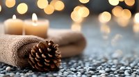 Cozy winter composition with pine cone, rolled towel and glowing candle against blurred golden bokeh lights creating warm atmospheric spa setting.