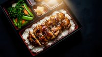 Japanese bento box with teriyaki chicken over rice, vegetables and sauce in traditional black lacquer container for authentic Asian cuisine dining.