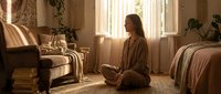 Young woman practicing mindfulness meditation in cozy bedroom with warm sunlight streaming through windows, sitting cross-legged on floor in comfortable clothing.