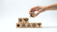 Hand placing wooden cube with gear symbol onto pyramid of blocks featuring various icons including eye, flower, sun, trinity knots, target, and caduceus on white background.