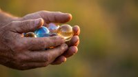 Close-up of weathered hands holding translucent glass spheres with blue and amber tones against blurred natural background. Suitable for concepts of care, fragility, and precious moments.
