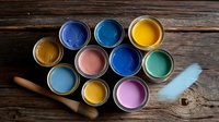 Paint cans with vibrant colors including yellow, blue, pink, and turquoise arranged on rustic wooden background with paintbrush, ready for home renovation project.
