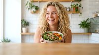 Happy blonde woman holding healthy salad bowl in modern kitchen with plants and wooden shelves for wellness lifestyle content.