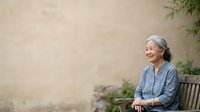 Senior Asian woman with gray hair sitting on wooden bench against neutral beige wall with green foliage, wearing blue shirt, contemplative pose for lifestyle and wellness content.