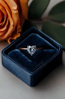 Heart-shaped diamond engagement ring in rose gold setting nestled in navy blue velvet ring box with orange rose petals, romantic proposal setup.